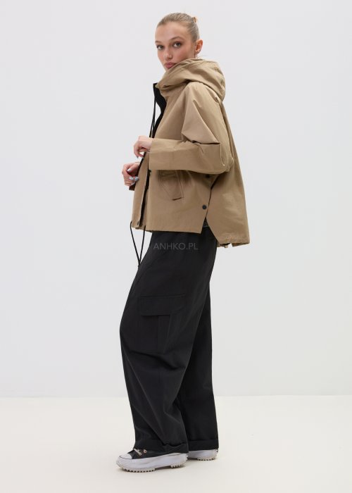 Camelowa kurtka damska z kapturem w stylu oversize, zapinana na napy, z regulacją w pasie i dużymi kieszeniami. Lekka kurtka o nowoczesnym kroju, zestawiona z czarnymi spodniami cargo.