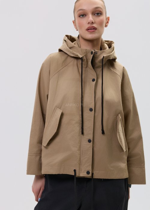 Camelowa kurtka damska z kapturem w stylu oversize, zapinana na napy, z regulacją w pasie i dużymi kieszeniami. Lekka kurtka o nowoczesnym kroju, zestawiona z czarnymi spodniami cargo.