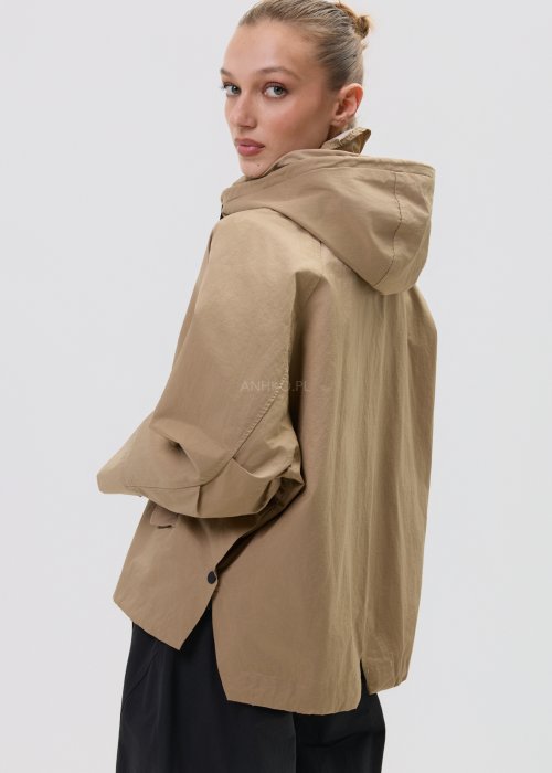 Camelowa kurtka damska z kapturem w stylu oversize, zapinana na napy, z regulacją w pasie i dużymi kieszeniami. Lekka kurtka o nowoczesnym kroju, zestawiona z czarnymi spodniami cargo.
