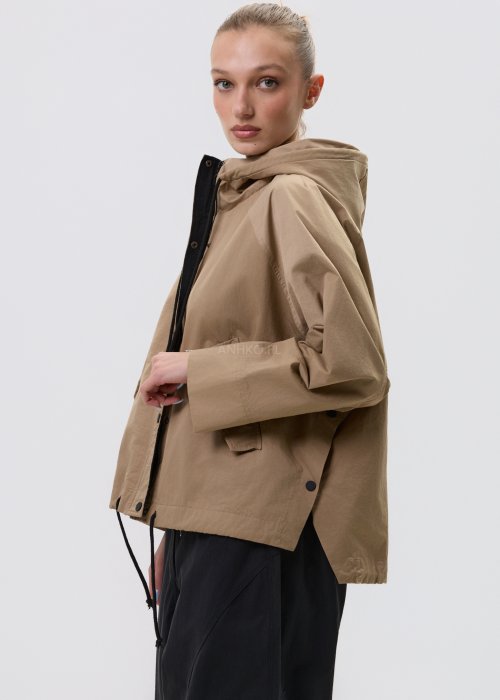 Camelowa kurtka damska z kapturem w stylu oversize, zapinana na napy, z regulacją w pasie i dużymi kieszeniami. Lekka kurtka o nowoczesnym kroju, zestawiona z czarnymi spodniami cargo.