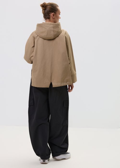 Camelowa kurtka damska z kapturem w stylu oversize, zapinana na napy, z regulacją w pasie i dużymi kieszeniami. Lekka kurtka o nowoczesnym kroju, zestawiona z czarnymi spodniami cargo.
