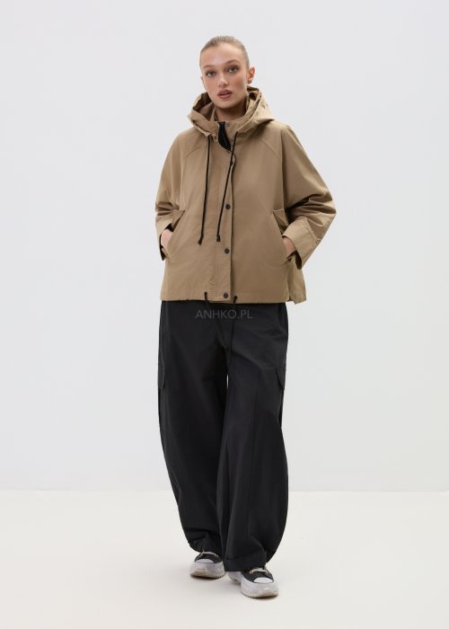 Camelowa kurtka damska z kapturem w stylu oversize, zapinana na napy, z regulacją w pasie i dużymi kieszeniami. Lekka kurtka o nowoczesnym kroju, zestawiona z czarnymi spodniami cargo.