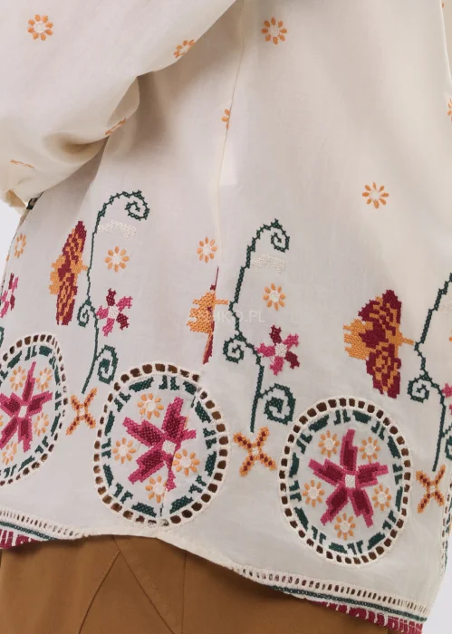 Kremowa koszula damska z delikatnym haftem kwiatowym i kolorową bordiurą u dołu, o lekkim, przewiewnym materiale i klasycznym kołnierzyku.