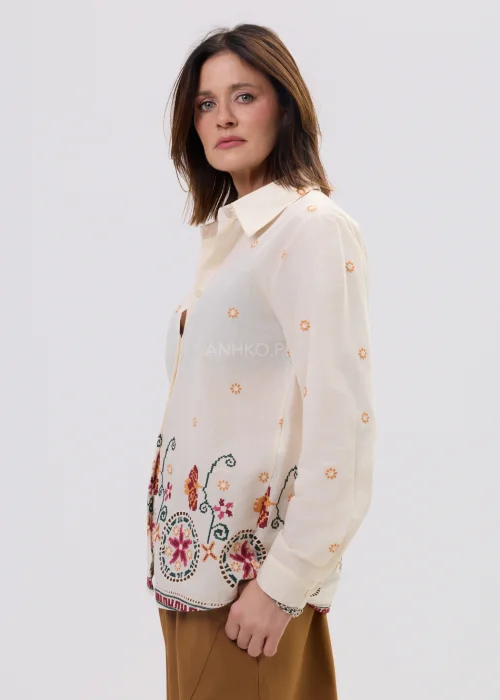 Kremowa koszula damska z delikatnym haftem kwiatowym i kolorową bordiurą u dołu, o lekkim, przewiewnym materiale i klasycznym kołnierzyku.