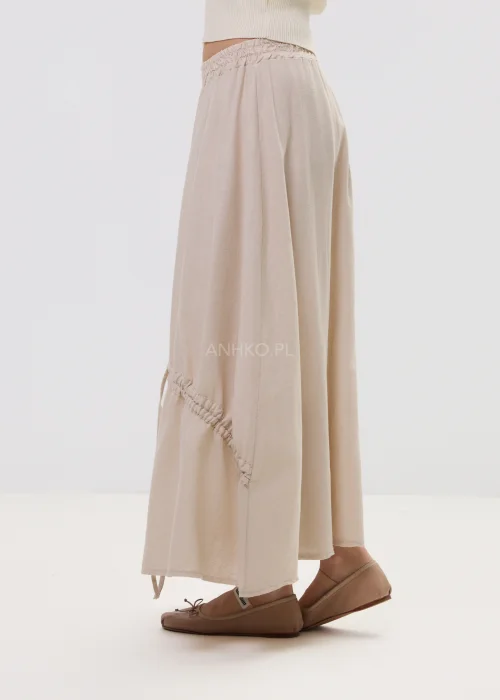 Beżowa spódnica maxi z elastyczną talią i dekoracyjnymi marszczeniami, lekki materiał, asymetryczne przeszycia – stylizacja z balerinami.
