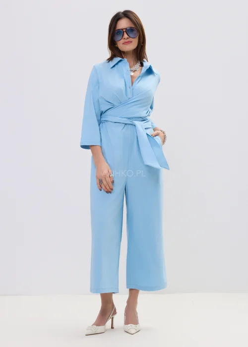 Niebieski kombinezon damski z kopertowym dekoltem i wiązaniem w talii, szerokie nogawki, elegancka stylizacja z szpilkami.