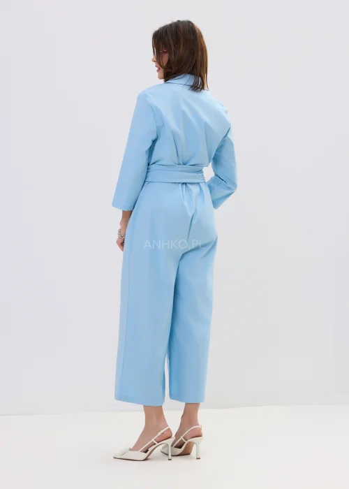 Niebieski kombinezon damski z kopertowym dekoltem i wiązaniem w talii, szerokie nogawki, elegancka stylizacja z szpilkami.