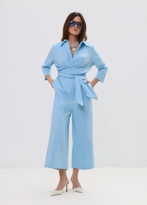 Niebieski kombinezon damski z kopertowym dekoltem i wiązaniem w talii, szerokie nogawki, elegancka stylizacja z szpilkami.