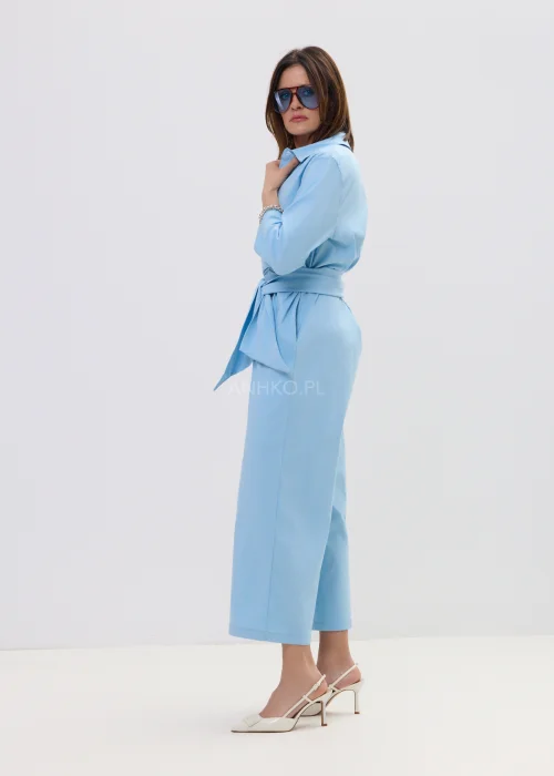 Niebieski kombinezon damski z kopertowym dekoltem i wiązaniem w talii, szerokie nogawki, elegancka stylizacja z szpilkami.