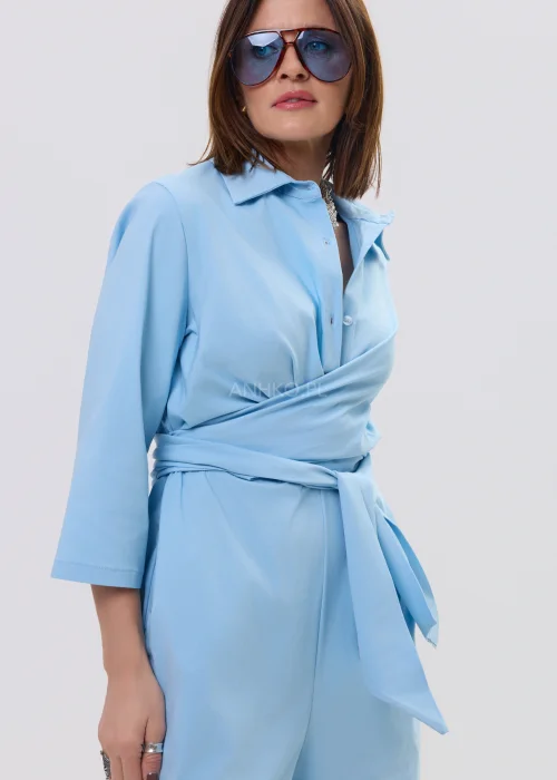 Niebieski kombinezon damski z kopertowym dekoltem i wiązaniem w talii, szerokie nogawki, elegancka stylizacja z szpilkami.