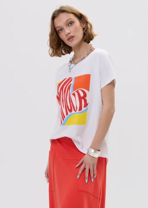 T-shirt oversize biały z kolorowym printem „Amour” w formie serca, luźny fason z krótkim rękawem – casualowa stylizacja z czerwono-pomarańczową spódnicą.
