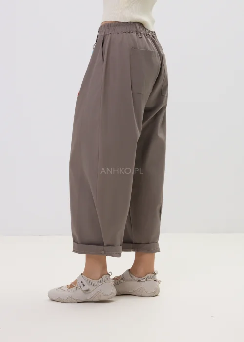 Spodnie damskie o luźnym kroju w kolorze taupe, z elastyczną talią i podwiniętymi nogawkami, ozdobione designerskim brelokiem – komfortowy model do codziennych stylizacji.