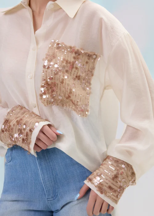 Beżowa koszula damska z cekinową kieszenią i mankietami, luźny fason, lekko transparentny materiał, styl casual-elegant.