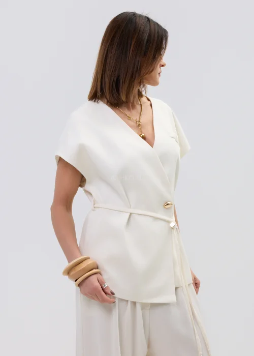 Śmietankowa kamizelka damska z dodatkiem lnu, kopertowy fason z paskiem w talii, elegancka stylizacja z jasnymi spodniami.