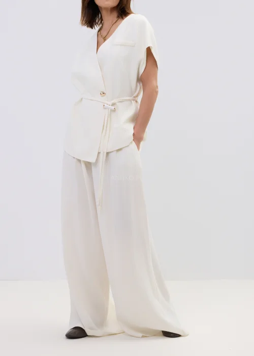 Śmietankowa kamizelka damska z dodatkiem lnu, kopertowy fason z paskiem w talii, elegancka stylizacja z jasnymi spodniami.