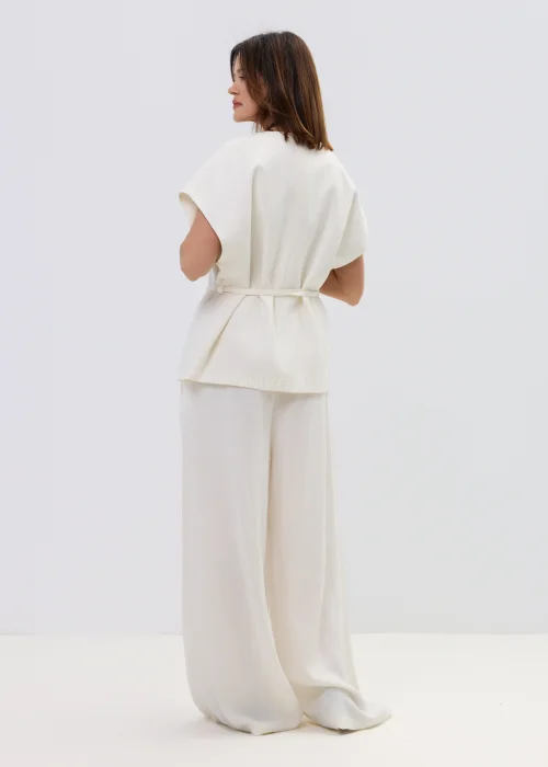 Śmietankowa kamizelka damska z dodatkiem lnu, kopertowy fason z paskiem w talii, elegancka stylizacja z jasnymi spodniami.