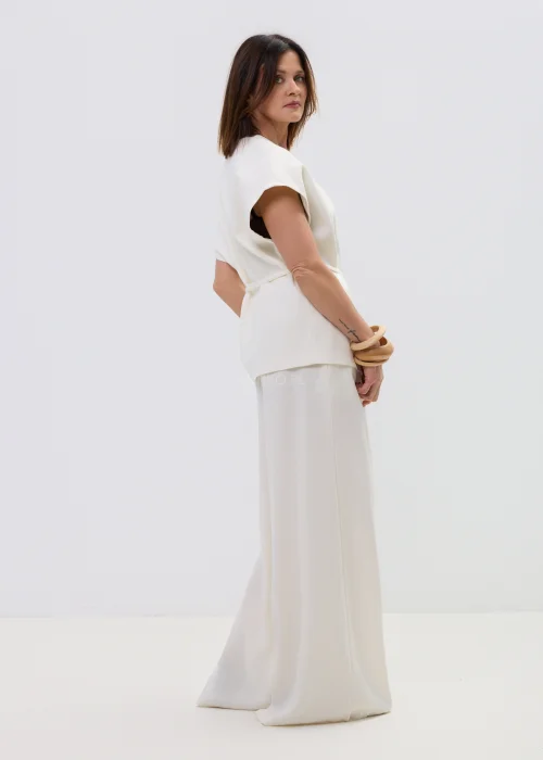 Śmietankowa kamizelka damska z dodatkiem lnu, kopertowy fason z paskiem w talii, elegancka stylizacja z jasnymi spodniami.