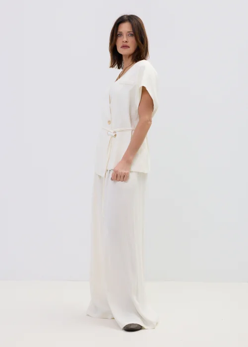 Śmietankowa kamizelka damska z dodatkiem lnu, kopertowy fason z paskiem w talii, elegancka stylizacja z jasnymi spodniami.