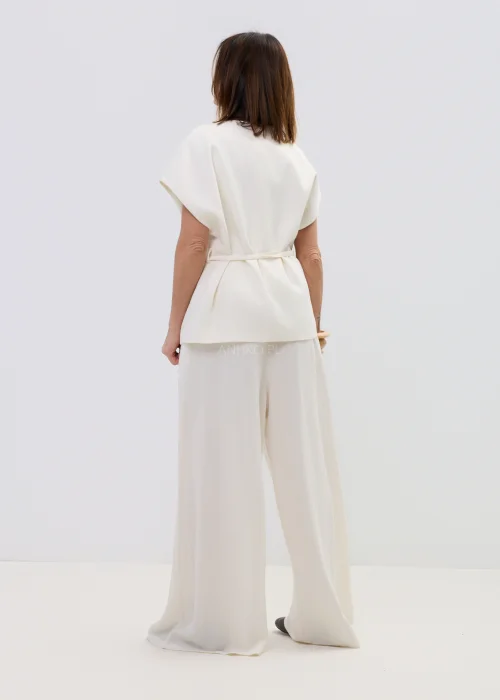 Śmietankowa kamizelka damska z dodatkiem lnu, kopertowy fason z paskiem w talii, elegancka stylizacja z jasnymi spodniami.