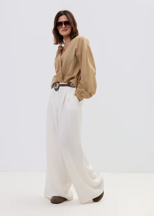 Camelowa bluzka damska z kopertowym dekoltem i falbaną, z długim rękawem, stylizowana z jasnymi spodniami i paskiem.