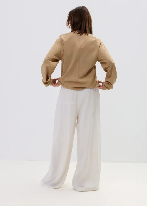 Camelowa bluzka damska z kopertowym dekoltem i falbaną, z długim rękawem, stylizowana z jasnymi spodniami i paskiem.
