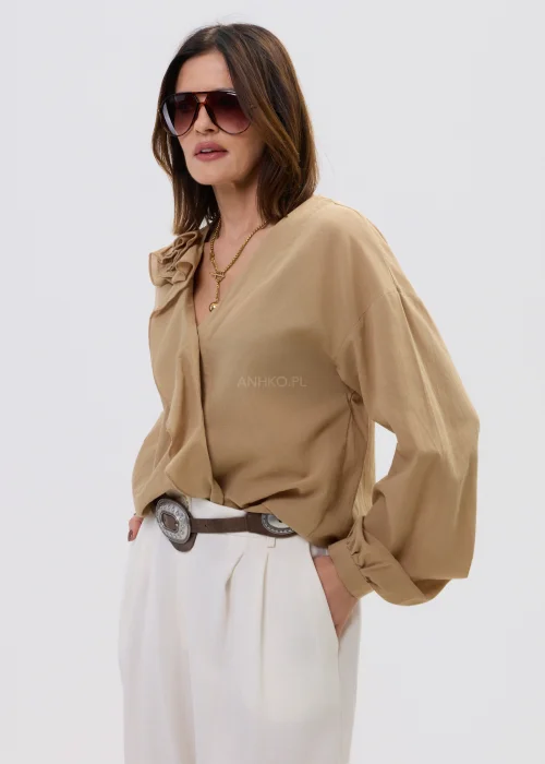 Camelowa bluzka damska z kopertowym dekoltem i falbaną, z długim rękawem, stylizowana z jasnymi spodniami i paskiem.