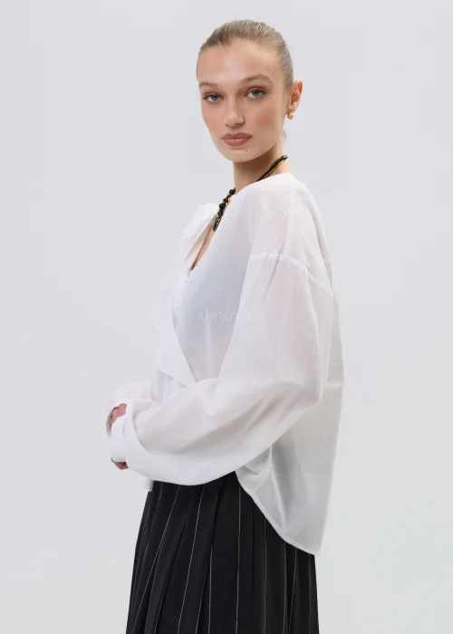 Biała bluzka kopertowa z delikatną falbaną i kwiatową aplikacją na ramieniu, z lekkiej półtransparentnej tkaniny, stylizowana z plisowaną spódnicą.