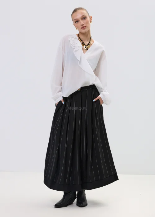 Biała bluzka kopertowa z delikatną falbaną i kwiatową aplikacją na ramieniu, z lekkiej półtransparentnej tkaniny, stylizowana z plisowaną spódnicą.