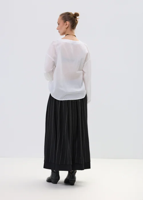 Biała bluzka kopertowa z delikatną falbaną i kwiatową aplikacją na ramieniu, z lekkiej półtransparentnej tkaniny, stylizowana z plisowaną spódnicą.