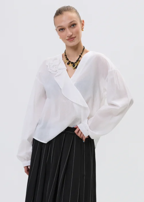 Biała bluzka kopertowa z delikatną falbaną i kwiatową aplikacją na ramieniu, z lekkiej półtransparentnej tkaniny, stylizowana z plisowaną spódnicą.