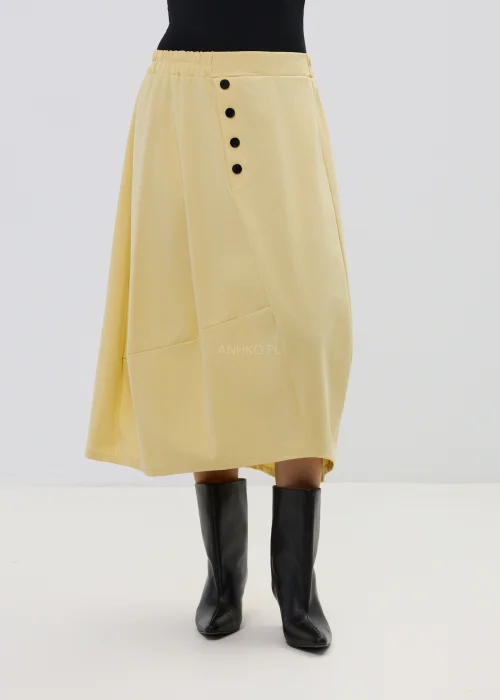 Żółta spódnica midi o asymetrycznym kroju z ozdobnymi czarnymi guzikami i elastyczną talią, elegancka i nowoczesna w stylizacji z wysokimi botkami.
