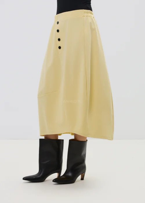 Żółta spódnica midi o asymetrycznym kroju z ozdobnymi czarnymi guzikami i elastyczną talią, elegancka i nowoczesna w stylizacji z wysokimi botkami.