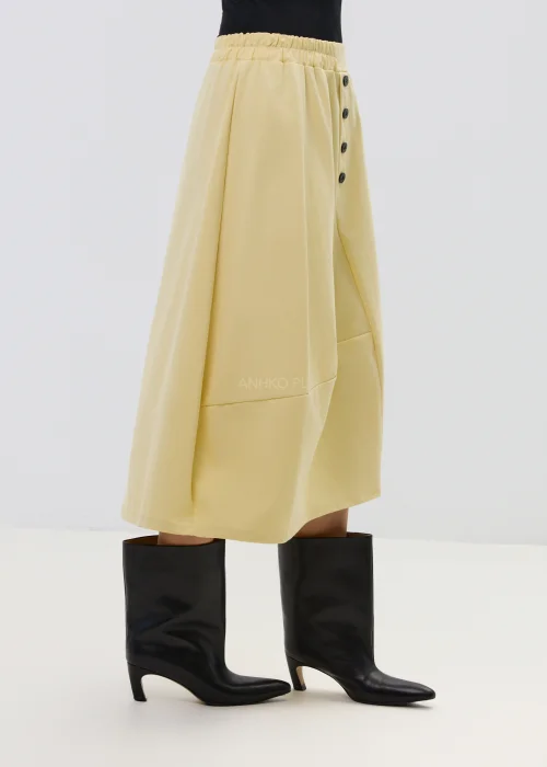 Żółta spódnica midi o asymetrycznym kroju z ozdobnymi czarnymi guzikami i elastyczną talią, elegancka i nowoczesna w stylizacji z wysokimi botkami.