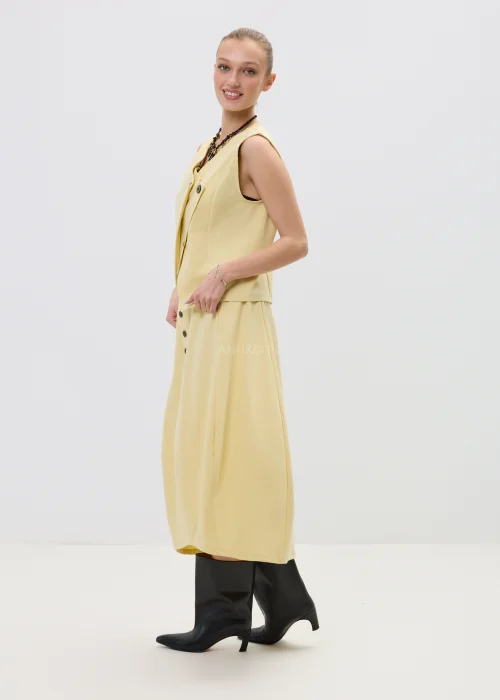 Żółta spódnica midi o asymetrycznym kroju z ozdobnymi czarnymi guzikami i elastyczną talią, elegancka i nowoczesna w stylizacji z wysokimi botkami.