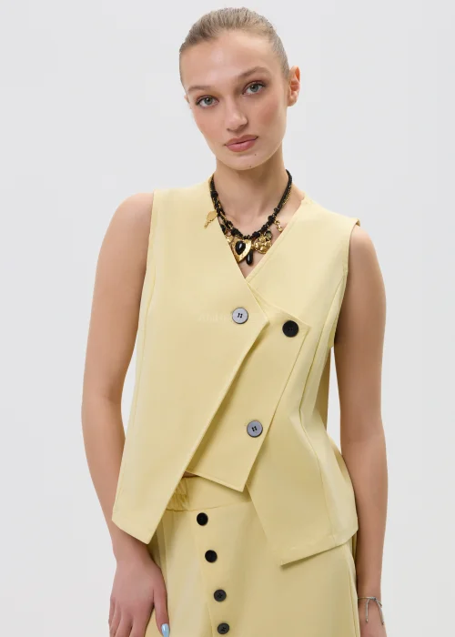 Żółta kamizelka damska o kopertowym kroju z ozdobnymi czarnymi guzikami, elegancka i nowoczesna, w stylizacji z dopasowaną spódnicą.