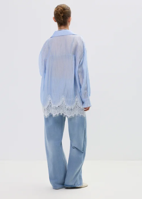 Niebieska koszula damska w delikatne paski z koronkowym wykończeniem u dołu, lekko transparentna, o luźnym kroju w stylizacji casual z jeansami.