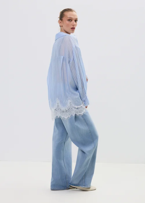 Niebieska koszula damska w delikatne paski z koronkowym wykończeniem u dołu, lekko transparentna, o luźnym kroju w stylizacji casual z jeansami.
