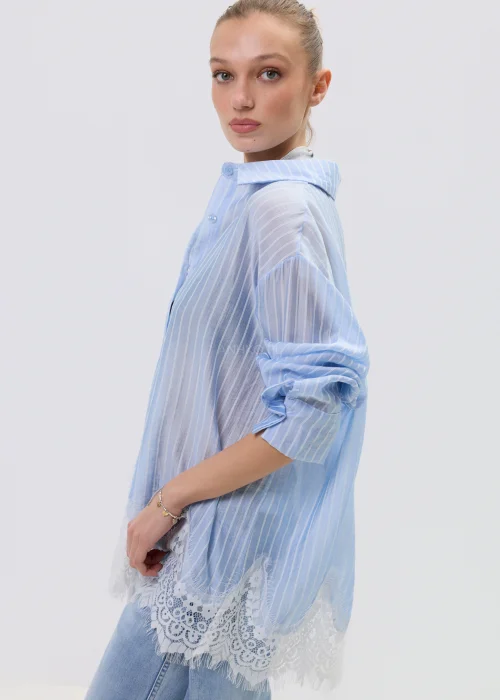 Niebieska koszula damska w delikatne paski z koronkowym wykończeniem u dołu, lekko transparentna, o luźnym kroju w stylizacji casual z jeansami.