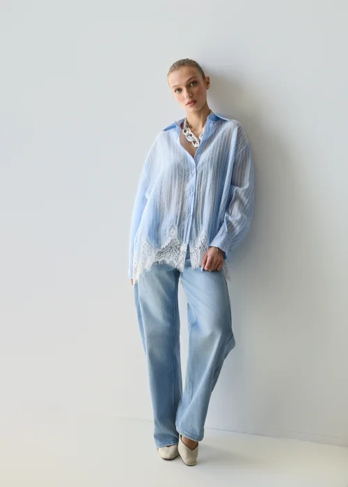 Niebieska koszula damska w delikatne paski z koronkowym wykończeniem u dołu, lekko transparentna, o luźnym kroju w stylizacji casual z jeansami.