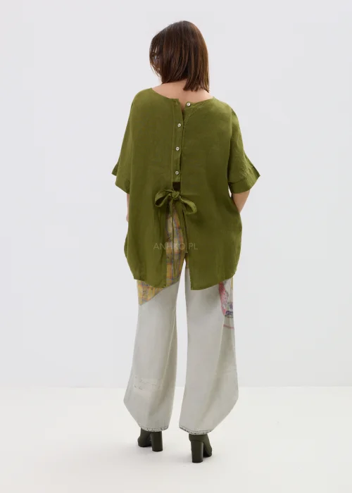 Lniana bluzka oversize w kolorze oliwkowej zieleni z krótkim rękawem i wiązaniem oraz guzikami na plecach, o luźnym kroju, stylizowana z jasnymi spodniami.