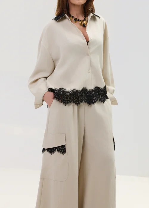 Koszula oversize w kolorze beżowym z czarną koronką przy kołnierzu i dole, o luźnym kroju z długim rękawem, stylizowana w elegancko-nowoczesnym zestawie.