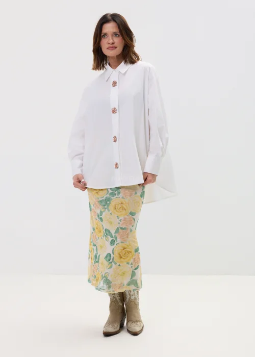 Koszula oversize w kolorze białym z ozdobnymi biżuteryjnymi guzikami w formie kwiatów, o luźnym kroju z szerokimi rękawami, stylizowana z kwiatową spódnicą.