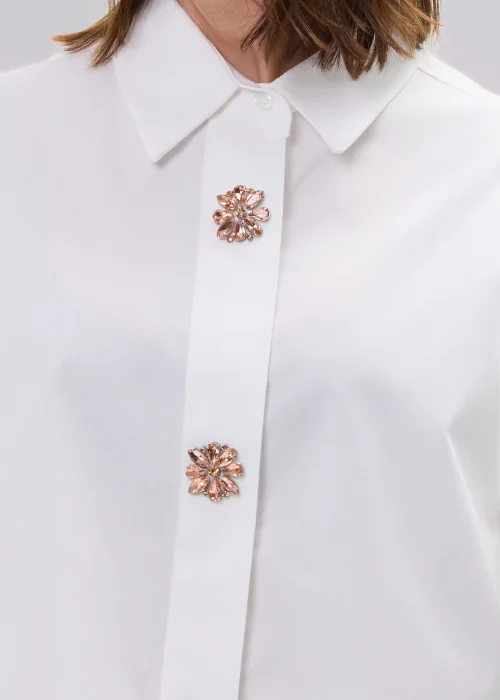 Koszula oversize w kolorze białym z ozdobnymi biżuteryjnymi guzikami w formie kwiatów, o luźnym kroju z szerokimi rękawami, stylizowana z kwiatową spódnicą.