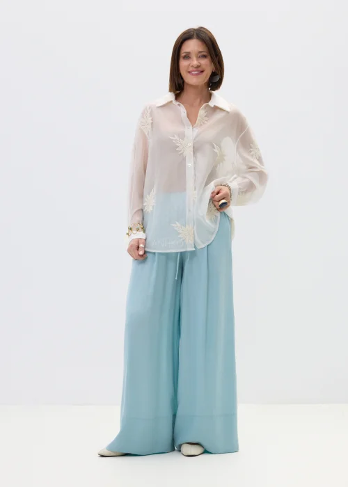 Transparentna biała koszula damska z delikatnym haftem kwiatowym, o luźnym kroju z długim rękawem i klasycznym kołnierzykiem, stylizacja warstwowa w lekkim, eleganckim klimacie.
