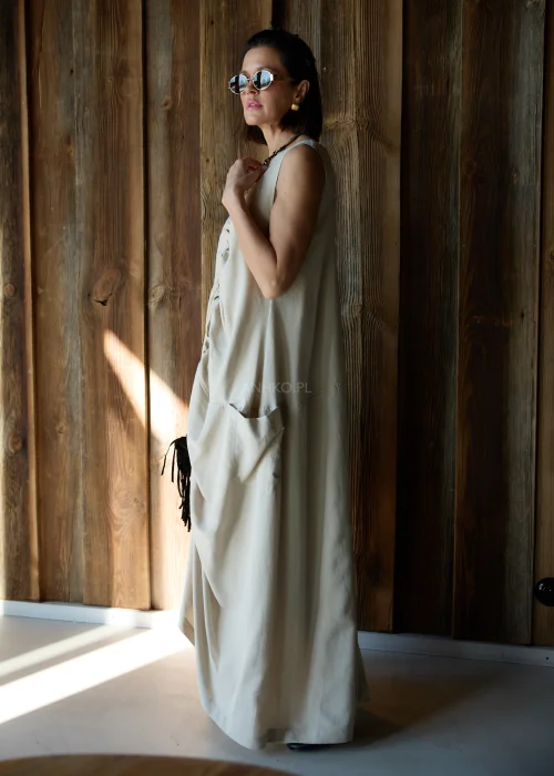 Beżowa sukienka maxi o luźnym kroju z marszczeniem z przodu i kieszeniami, stylizowana z okularami przeciwsłonecznymi i biżuterią.