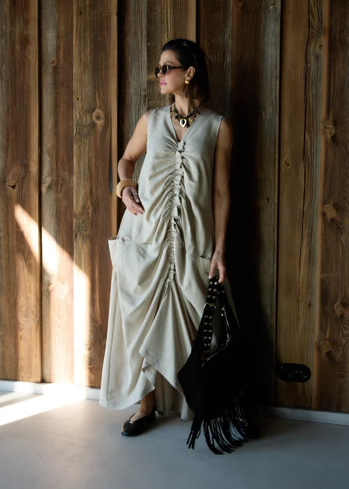 Beżowa sukienka maxi o luźnym kroju z marszczeniem z przodu i kieszeniami, stylizowana z okularami przeciwsłonecznymi i biżuterią.