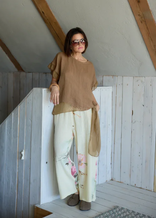 Lniana bluzka damska w jasnobrązowym kolorze z falbankami przy rękawach i wiązaniem na plecach, stylizacja z lekkimi spodniami w naturalnym stylu.