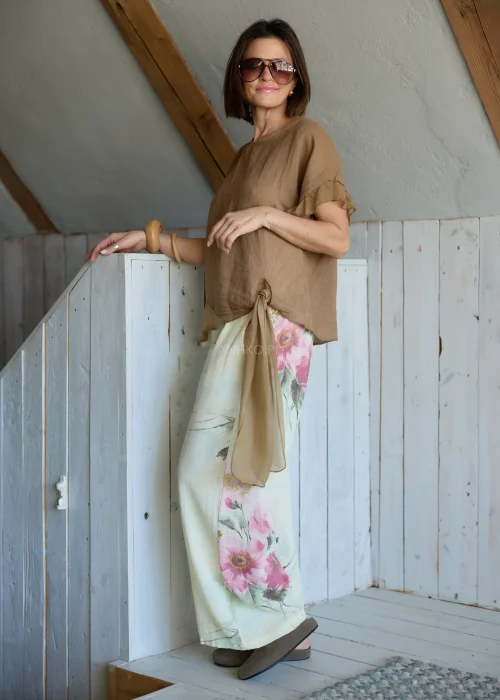 Lniana bluzka damska w jasnobrązowym kolorze z falbankami przy rękawach i wiązaniem na plecach, stylizacja z lekkimi spodniami w naturalnym stylu.