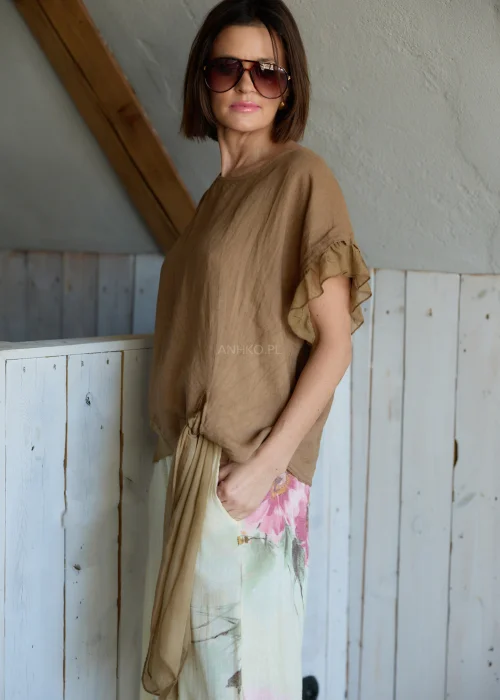 Lniana bluzka damska w jasnobrązowym kolorze z falbankami przy rękawach i wiązaniem na plecach, stylizacja z lekkimi spodniami w naturalnym stylu.