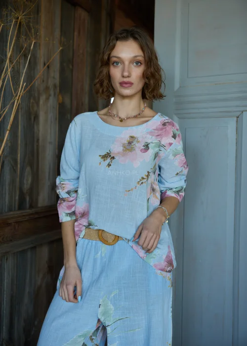 Bluzka damska w kolorze błękitnym z kwiatowym printem, o luźnym kroju, stylizowana z jasnymi spodniami i paskiem w naturalnym stylu.